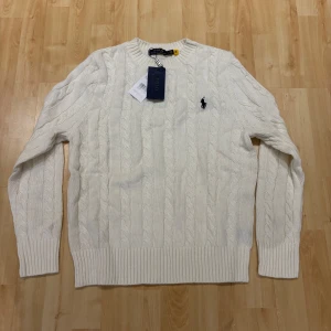 Vit kabelstickad tröja Polo Ralph Lauren - Vit kabelstickad tröja från Polo Ralph Lauren med klassisk rund halsringning och broderad logga på bröstet. Tröjan har ribbade muddar och är tillverkad i mjuk bomull. Perfekt för dig som gillar en clean och tidlös stil.