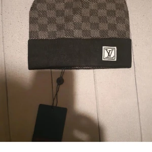 Rutig mössa från Louis Vuitton - Snygg mössa från Louis Vuitton med klassiskt rutigt mönster i svart och grått. Mössan har en bred svart uppvikt kant med LV-logga framtill. Perfekt accessoar för att lyfta din streetstyle. Materialet känns mjukt och bekvämt.