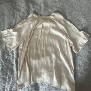 Ljusbeige blus i siden från Wera - Supersnygg ljusbeige blus från Wera i glansigt sidenmaterial. Blusen har korta, vida ärmar och en avslappnad passform med lätt rynk vid halsringningen. Perfekt att styla med jeans eller kjol för en clean look.