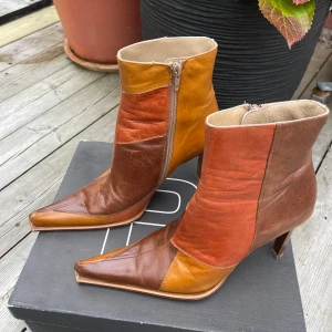 Patchwork stövletter från Bianco - Unika stövletter i patchwork-stil från Bianco med höga klackar och spetsig tå. Skorna är i skinn och har paneler i brunt, orange och senapsgult. Dragkedja på insidan för enkel påtagning. Perfekta för dig som vill sticka ut med retrovibbar. Köpta på 90-talet. 
