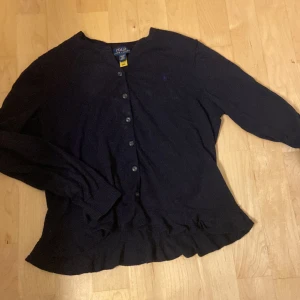Mörkblå kofta från Polo Ralph Lauren - Snygg mörkblå kofta från Polo Ralph Lauren i klassisk modell med knappar framtill. Långärmad och med ribbade muddar samt volangdetalj nedtill. 