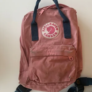 Rosa Kånken Mini ryggsäck från Fjällräven - Säljer en rosa Kånken Mini ryggsäck från Fjällräven med mörkblå axelremmar och handtag. Klassisk fyrkantig form, stort huvudfack med dragkedja och en mindre ficka framtill. Tillverkad i slitstark polyester och har det ikoniska runda Kånken-märket på framsidan.