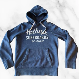 Blå hoodie från Hollister med tryck - Mysig blå hoodie från Hollister med broderat vitt tryck 'Hollister Surfboards SO.CALIF' på bröstet. Tröjan har huva med vita snören och en stor magficka. Perfekt för chill dagar och snygg till jeans eller joggers.