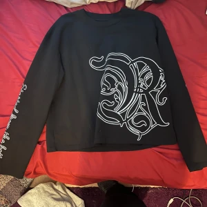 Svart sweatshirt Rise Up med tryck - Svart långärmad sweatshirt från Rise Up med stora vita tryck fram och bak. Framsidan har en stor dekorativ bokstav, baksidan texten 'seen this angel?' och ärmen har texten 'on my way to the top'. Skön passform och cool streetstil.