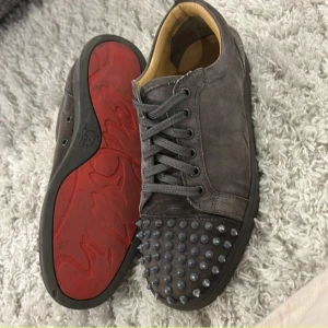 Gråa Louboutin sneakers med nitar - Säljer ett par gråa sneakers från Christian Louboutin i mocka med ikonisk röd sula och coola nitar på tån. Skorna har snörning och en rund tå, perfekt för dig som vill sticka ut med en edgy vibe. Insidan är beige och sulan är svart med röd undersida.