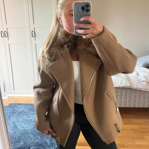 Beige pälsjacka med dragkedjor - Snygg beige jacka med bred krage i fuskpäls och coola silvriga dragkedjor framtill. Jackan har en loose passform och stora slag, perfekt för dig som gillar statement-plagg. Materialet känns tjockt och mjukt, och pälsdetaljen ger extra edge till looken. Det ska finnas ett band längst ner som tyvärr är borta men använt jacka ändå och man tänker inte på det