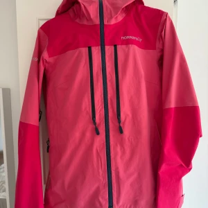 Rosa skaljacka från Norrøna med huva - Snygg tvåfärgad rosa vindjacka från Norrøna med huva och svarta dragkedjor framtill. Jackan har långa ärmar, justerbara muddar och praktiska fickor med dragkedja. Perfekt för utomhusaktiviteter och äventyr.