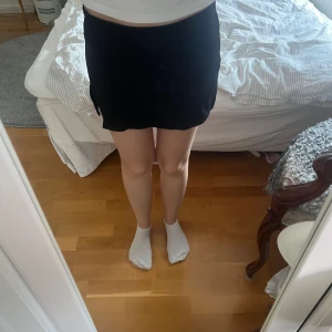 Svart kortkjol med slits från Monki - Svart kortkjol från Monki i stretchigt material. Kjolen har en enkel design med en liten slits på sidan som ger en snygg detalj. Perfekt att matcha med en crop top eller oversized hoodie. Midjehög passform och mjuk känsla.