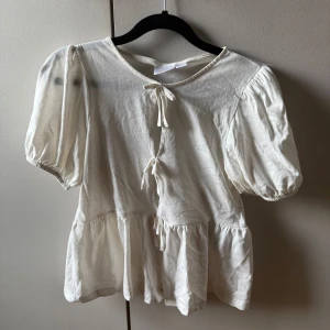 Vit blus med knyt från Gina Tricot - Supersöt vit blus från Gina Tricot med puffiga korta ärmar och volang nertill. Blusen har tre knyt framtill som ger en gullig och trendig look. Tillverkad i mjuk bomull, perfekt för dig som gillar romantisk stil.