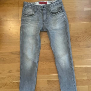 Grå Replay Anbass - Säljer ett par grå jeans från Replay med snyggt slitna detaljer och klassisk femficksdesign. Jeansen har svarta sömmar, Replay-logga på fickan och bak. Perfekta för dig som gillar en modern och avslappnad look. Modell anbass. 28/32.