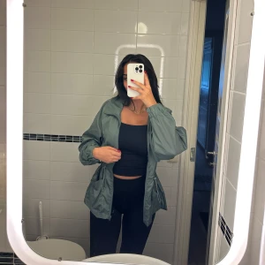 Grön vindjacka med snörning - Snygg grön vindjacka med dragkedja framtill och snörning i midjan. Jackan har långa ärmar med resår vid handlederna och praktiska fickor på sidorna. Perfekt för dig som gillar en avslappnad och trendig stil.