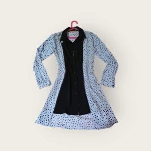 Prickig skjortklänning i svart och vitt - Snygg skjortklänning med svart mittparti och vita sidor med svarta prickar. Långärmad modell med knappar framtill och klassisk krage. Klänningen har en lätt utställd kjol och är knälång. Perfekt för dig som gillar kontraster och lekfulla mönster.