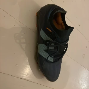 Svarta New Balance fotbollsskor - Säljer ett par svarta New Balance fotbollsskor med stora ljusgrå N-loggor på sidorna. Skorna har snörning och är designade för att ge bra grepp på planen. Snygg och modern look med orange detaljer på sulan och insidan. Pris går att diskuteras