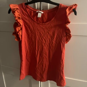 Röd volangtopp från H&M - Snygg röd topp från H&M  med volangdetaljer på axlarna. Modellen är ärmlös och har en rund halsringning. Perfekt för dig som vill ha en enkel men ändå trendig look.