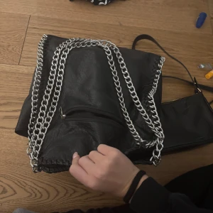 Tiamo Svart axelväska med kedja  - Snygg svart axelväska i skinnimitation med grova silverfärgade kedjor längs kanterna och som axelrem. Väskan har en dragkedja framtill och en cool, lite slouchy form. Perfekt för dig som gillar edgy accessoarer och vill sticka ut.