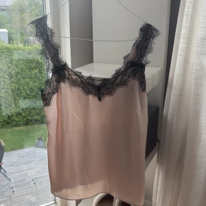 Ljusrosa linnetopp med svart spets - Säljer en ljusrosa linnetopp med breda axelband och vacker svart spets längs urringningen. Toppen har en slät, glansig yta och är ärmlös, perfekt för att skapa en chic och feminin look.