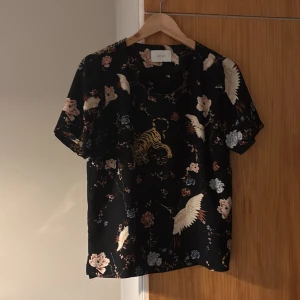 Svart blommig blus från Neo Noir - Svart blus från Neo Noir med korta ärmar och ett snyggt blommigt mönster, samt detaljer med tigrar och tranor. Skön passform och lätt material, perfekt för dig som vill sticka ut lite extra.