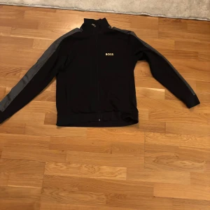 Svart ziptröja från BOSS med gråa detaljer - Svart ziptröja från BOSS med gråa ränder längs ärmarna och guldfärgad BOSS-logga på bröstet. Hög krage och hel dragkedja framtill. Perfekt för en sportig och clean look. Ny pris 1200