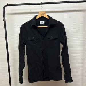 Svart Overshirt från Ciszere - Svart Overshirt från Ciszere med klassisk krage och två bröstfickor med lock och knappar. Skjortan har lång ärm och stängs med knappar framtill. Snygg och stilren modell som passar till många olika looks.