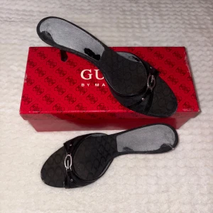 Svarta sandaler från Guess med logga - Snygga svarta sandaler från Guess med öppen tå och låg klack. Skorna har ett diskret mönster med Guess-logga på hela ovandelen och en silverfärgad detalj framtill. Tillverkade i syntetmaterial med läderdetaljer och levereras med originalkartong och dustbag.