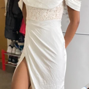 Vit långklänning med spets och slits - Snygg vit långklänning med offshoulder och spetsdetaljer upptill. Klänningen har en hög slits på ena sidan och är figurnära med draperad överdel. Priset kan diskuteras 