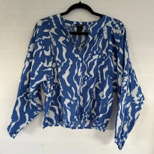 Blåvit blus med mönster - Säljer en blå och vit blus från Lindex i storlek XS. Blusen har ett unikt abstrakt mönster, v-ringning och långa ärmar. Tillverkad i tunn bomull, perfekt för dig som gillar statement-plagg med cool design.🤩💕