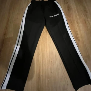 Palm Angels svarta trackpants med vita ränder - Svarta trackpants från Palm Angels med vita sidoränder och logga tryckt på vänster lår. Byxorna har elastisk midja och raka ben, perfekt för en sportig och trendig streetwear-look.
