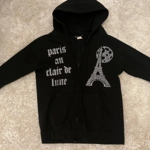 Svart hoodie med strass och Paris-tryck - Svart hoodie från Blingin med dragkedja, huva och fickor. Framsidan har strassdetaljer med texten 'paris au clair de lune' och motiv av Eiffeltornet och månen. Perfekt statement-plagg för dig som gillar Paris-vibes och bling, säljer då den är för liten!