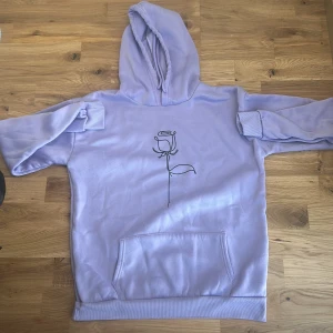 Ljuslila hoodie med ros-tryck XL 170 92A - Ljuslila hoodie i storlek XL med en stilren svart linjeteckning av en ros på bröstet. Tröjan har huva med dragsko, magficka och ribbade muddar vid ärmslut och nederkant. Perfekt för dig som gillar en chill och snygg stil.