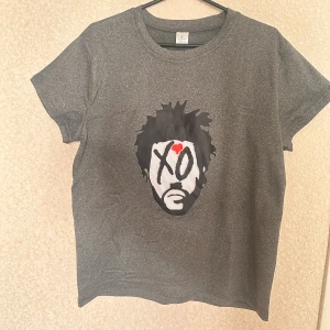 The weeknd XO t-shirt grå - Säljer en grå t-shirt med ett stort grafiskt tryck av ett ansikte och texten XO på bröstet. Trycket har svart, vitt och rött i designen. T-shirten har rund hals och korta ärmar, perfekt för dig som gillar streetwear och coola prints.