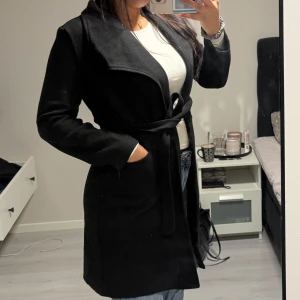 Svart kappa med bälte från Shein - Stilren svart kappa från Shein med knytskärp i midjan och stora fickor. Jackan har en klassisk krage och är långärmad, perfekt för dig som gillar en clean och enkel look. Materialet är mjukt och känns skönt mot kroppen.