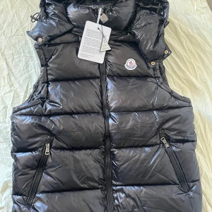 Moncler Väst - Svart glansig dunväst från Moncler med huva och dragkedja framtill. Storleken är M men passformen är mer som S. Västen har två snedställda fickor med dragkedja och ikonisk Moncler-logga på bröstet. Insidan har ett coolt serietryck och autenticitetsmärkning. Perfekt för lager-på-lager under kyliga dagar.