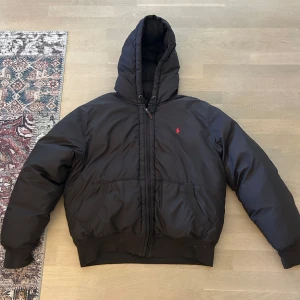 Vintage pufferjacka Polo Ralph Lauren - Svart pufferjacka från Polo Ralph Lauren med huva och dragkedja. Jackan har två stora fickor framtill och ikonisk röd Polo-logga broderad på bröstet. Fodrad och riktigt varm, perfekt för kalla dagar. Klassisk och stilren design med ribbade muddar.