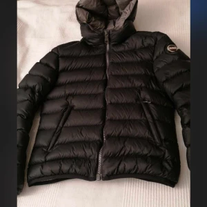 Svart dunjacka från Colmar, strl 44 - Snygg svart dunjacka från Colmar med grå insida och huva. Jackan har quiltad design, två snedställda fickor med dragkedja och tydlig logga på ärmen. Perfekt för kalla dagar och har en modern, sportig look. Strl 44 sitter som en S/M