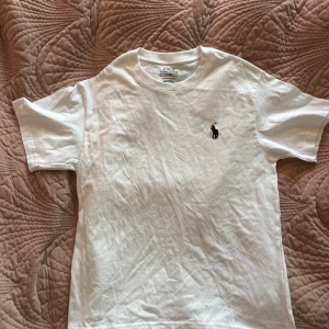 Vit t-shirt från Polo Ralph Lauren - Vit t-shirt från Polo Ralph Lauren med klassisk svart broderad logga på bröstet. Modellen är custom slim fit med rund hals och korta ärmar. Tillverkad i mjuk bomull, perfekt för en clean och stilren look.