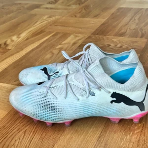Puma vita fotbollsskor med rosa dobbar - Säljer ett par snygga fotbollsskor från Puma i vitt med svarta loggor och coola rosa dobbar undertill. Skorna har snörning och är tillverkade i syntetmaterial med stickad känsla upptill. Perfekta för dig som vill sticka ut på planen!