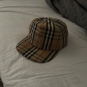 Burberry rutig keps i beige och svart - Snygg keps från Burberry med klassiskt rutigt mönster i beige, svart, vitt och röda detaljer. Märket syns tydligt baktill med texten 'Burberry London England'. Kepsen är tillverkad i bomull och polyester och har justerbar rem baktill för perfekt passform.