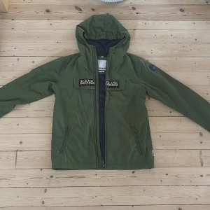 Grön vindjacka från Napapijri - Snygg grön vindjacka från Napapijri med huva och dragkedja framtill. Jackan har två fickor med dragkedja, broderad logga på bröstet och patch på ärmen. Materialet är polyester och insidan har meshfoder. Perfekt för blåsiga dagar.
