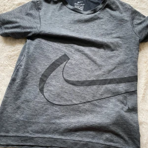 Grå Nike Dri-Fit t-shirt med stor logga - Grå Nike Dri-Fit t-shirt med stor svart swoosh-logga framtill. T-shirten är kortärmad och tillverkad i ett mjukt, funktionsmaterial som andas. Perfekt för träning eller sportiga dagar. Enkel och stilren design med rund hals.