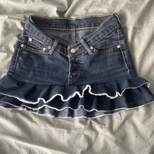 Levi's jeanskjol med volangkant - Supersöt jeanskjol från Levi's i klassisk blå denim med volangkant nertill och kontrastsömmar. Kjolen har fem fickoroch tre knappar framtill samt vita detaljer längs volangen. Perfekt för dig som vill ha en trendig och lekfull look.