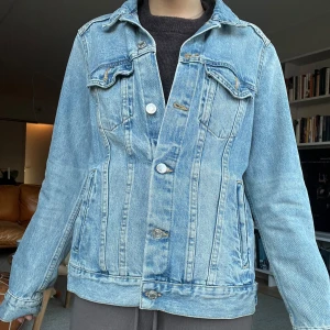 Ljusblå jeansjacka med knappar - Klassisk ljusblå jeansjacka med raka linjer och silverfärgade knappar framtill. Jackan har två bröstfickor med lock och knapp, samt två sidofickor. Snygg krage och långärmad modell i ett slitstarkt denimtyg. Perfekt för lager-på-lager-stil.