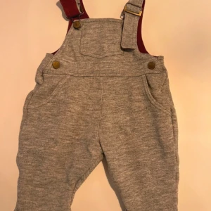 Grå mjukisbyxdress med röda hängslen - Grå mjukisbyxdress i mjukt material med röda axelband och mässingsfärgade knappar. Byxdressen har fickor fram och bak samt justerbara axelband. Perfekt för en chill och avslappnad stil. Storlek 62/68