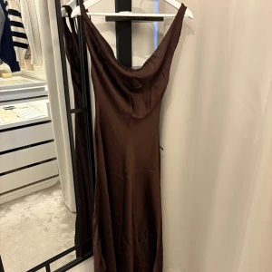 Oanvänd brun långklänning i satin från Zara - Supersnygg brun långklänning i satin från Zara med draperad halsringning och öppen rygg med knyt. Klänningen är ärmlös och har en elegant, figurnära passform som faller snyggt längs kroppen. Perfekt för dig som vill sticka ut med en stilren look. 