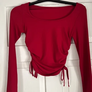 Röd  långärmad topp - Snygg röd långärmad topp med ribbad struktur och trendig snörning i sidorna som ger en cool rynkad effekt. Modellen är croppad och har en rundad halsringning. Perfekt för dig som vill sticka ut med färg och detaljer.