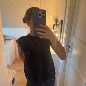 Mörkblå spetsblus med volang - Säljer en mörkblå blus i spets med blommigt mönster och volangdetaljer längs axlar och framstycke. Blusen är ärmlös med hög hals och genombrutet tyg.