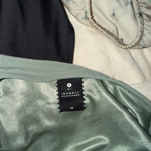 Grön omlottopp från Isabell Kristensen - Säljer en ljusgrön omlottopp från Isabell Kristensen med broderade detaljer vid v-ringningen och smockad midja. Toppen är ärmlös och har en mjuk, glansig yta som känns lyxig. Perfekt att styla med jeans för en chill look.