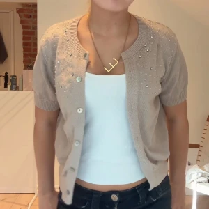 Beige kofta med pärlor och kort ärm - Supersöt beige kofta med korta ärmar och pärldetaljer upptill. Koftan har rund halsringning och knäpps med knappar framtill. Perfekt att slänga över ett linne för en chill och snygg look. Knappt använd