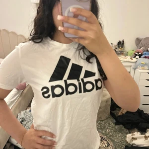Vit Adidas t-shirt - Vit t-shirt från Adidas med stor svart logga framtill. Klassisk rund hals och korta ärmar. Skön passform och tillverkad i mjuk bomull, perfekt för en chill och sportig look. passar både storlek XS och S till och med mindre kanske.