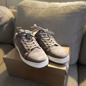 Gråa sneakers från Christian Louboutin - Snygga gråa sneakers från Christian Louboutin med klassisk röd sula. Skorna har snörning och är tillverkade i mocka med vita kanter runt sulan. Perfekta för dig som vill ha en stilren och exklusiv look med en touch av lyx.