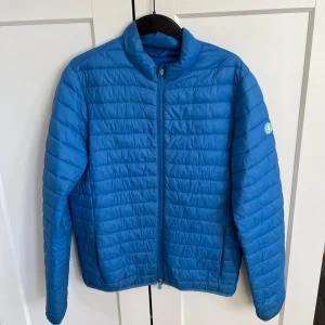 Blå quiltad pufferjacka med dragkedja - Säljer en blå pufferjacka från Save the Duck med quiltad design och hög krage. Jackan har lång ärm, två sidofickor och hel dragkedja framtill. Märkets logga finns på vänster ärm. Perfekt för dig som gillar stilren och sportig look. Tyvärr saknas dragkedjans del som man drar ner med. Men går att sätta i ny o hitta för billigt pris